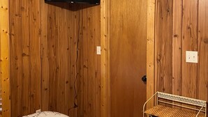 Quarto com paredes de madeira, ventilador de teto, televisão de tela plana acima de uma porta, cama com colcha estampada e cesta de vime para guardar objetos.