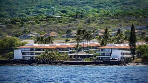Un hôtel situé sur la côte, composé de plusieurs bâtiments, dont certains sont surmontés de toits rouges, et dont l’arrière-plan est constitué d’une végétation luxuriante et de collines.