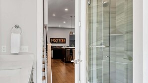 Ein modernes Badezimmer mit Duschkabine aus Glas, einem weißen Waschbecken und einem sichtbaren Küchenbereich im Hintergrund.