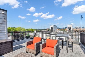 Une terrasse extérieure avec des meubles en osier et des coussins orange, une clôture en métal noir et une vue sur la ville en arrière-plan.