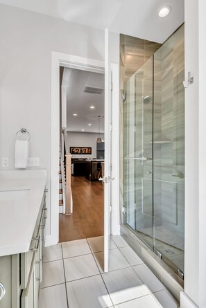 Une salle de bain moderne avec une douche à parois de verre, un lavabo blanc et une cuisine visible à l’arrière-plan.