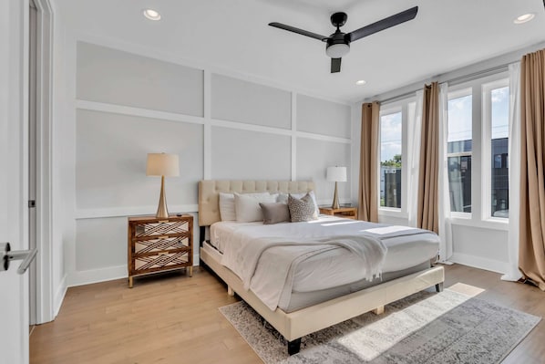 A bedroom with a large bed, a ceiling fan, and a view of the outside through large windows.