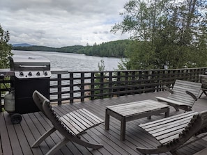 Una terrazza con mobili in legno e un barbecue, con vista su un lago, alberi e colline sullo sfondo.