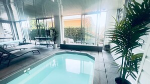 Ein Hallenbad mit großen Glaswänden, eine Lounge-Bereich mit Sesseln und einem Tisch sowie Kübelpflanzen.