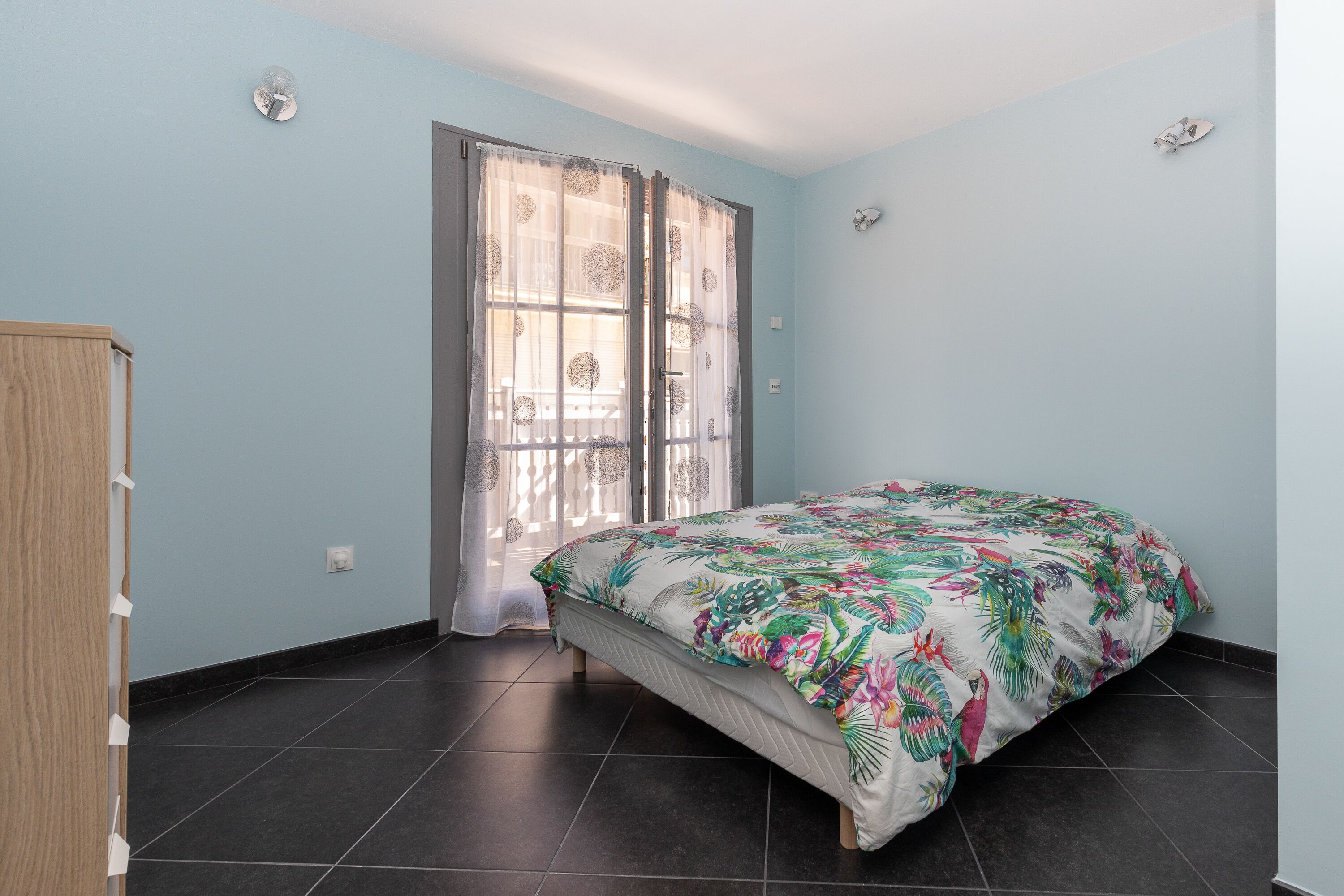A bedroom with a bed, a wardrobe, a window with curtains, and a tiled floor.