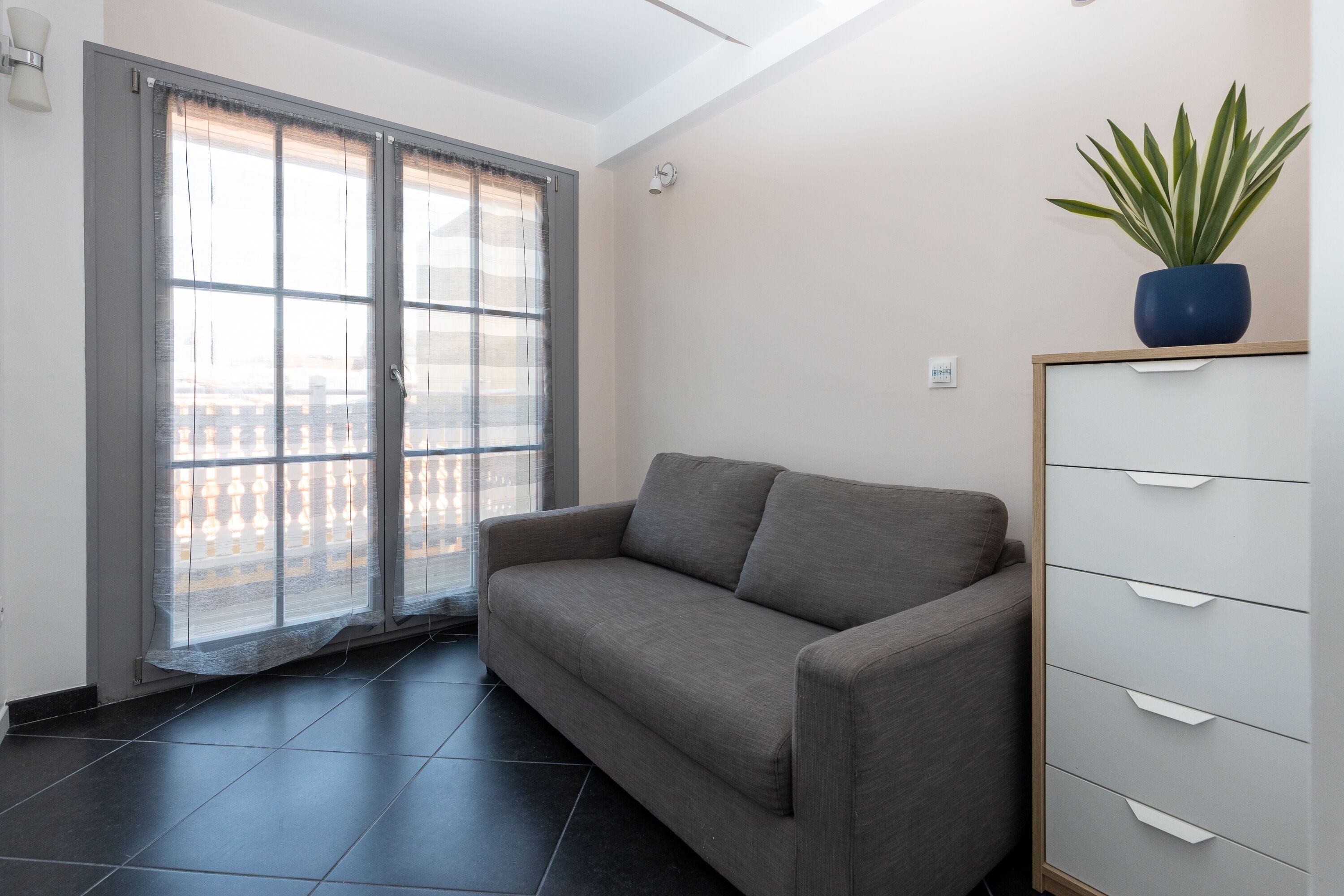 Una sala de estar con un sofá gris, un mueble con cajones, una planta en maceta y vistas a través de una ventana con cortina.