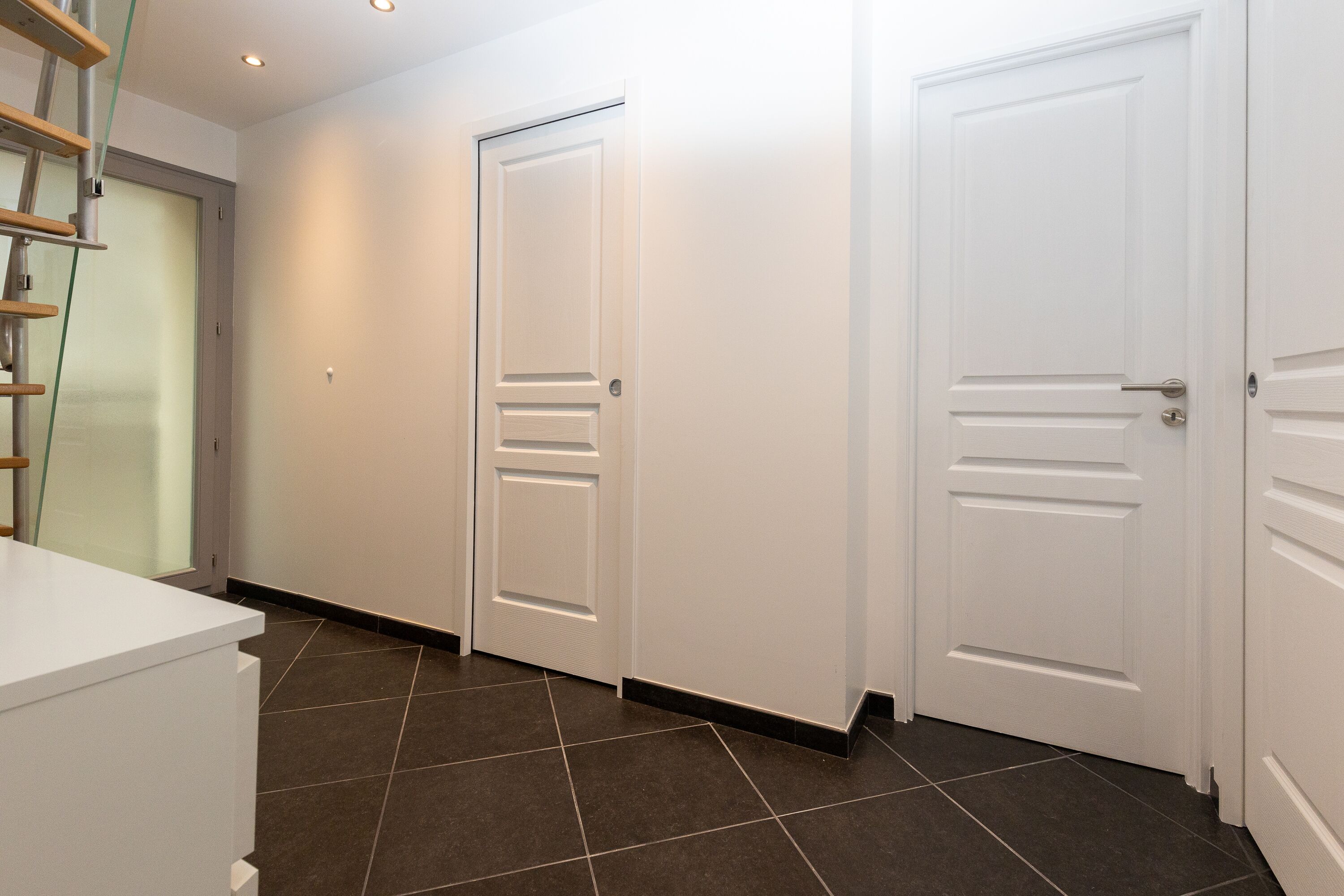 A hallway with three white doors, a white counter, and dark tiled flooring.