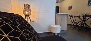 Un salon moderne avec un canapé à motifs géométriques, un petit tabouret rond, une table basse en verre, une lampe au design original en fil de métal, un meuble en bois et un coin repas avec une table et des chaises.