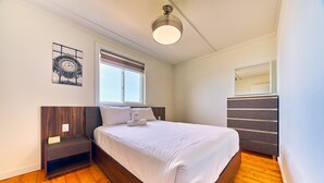 A bedroom with a wooden floor, a bed with white bedding, a nightstand, a dresser, a window with blinds, and a wall-mounted mirror. - Suite 201: Beautiful 3-Bedroom Beachfront Townhouse (Wainfleet)