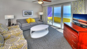 Ein Hotelzimmer mit grauem Sofa, gelben Kissen, einem weißen Couchtisch, einem roten Schminktisch, einem Deckenventilator, einem Fernseher und einem Balkon mit Meerblick.