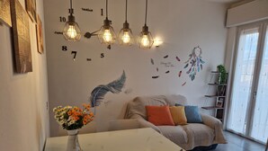 Ein modernes Wohnzimmer mit einem weißen Sofa, farbenfrohen Kissen, einem Esstisch mit Stühlen und einer Wanduhr.