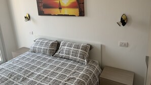 Ein Schlafzimmer mit einem Bett, gestreiften Kissen, einem Wandbild mit Sonnenuntergang über Wasser und zwei Wandleuchten.