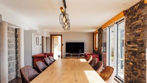 Un espace repas moderne avec une longue table en bois, des chaises en cuir, un téléviseur encastré et un mur d’accent en pierre.