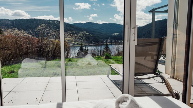 Une pièce dotée d’une grande fenêtre en verre donnant sur un lac et des montagnes. On y trouve une chaise et une serviette sur le lit.