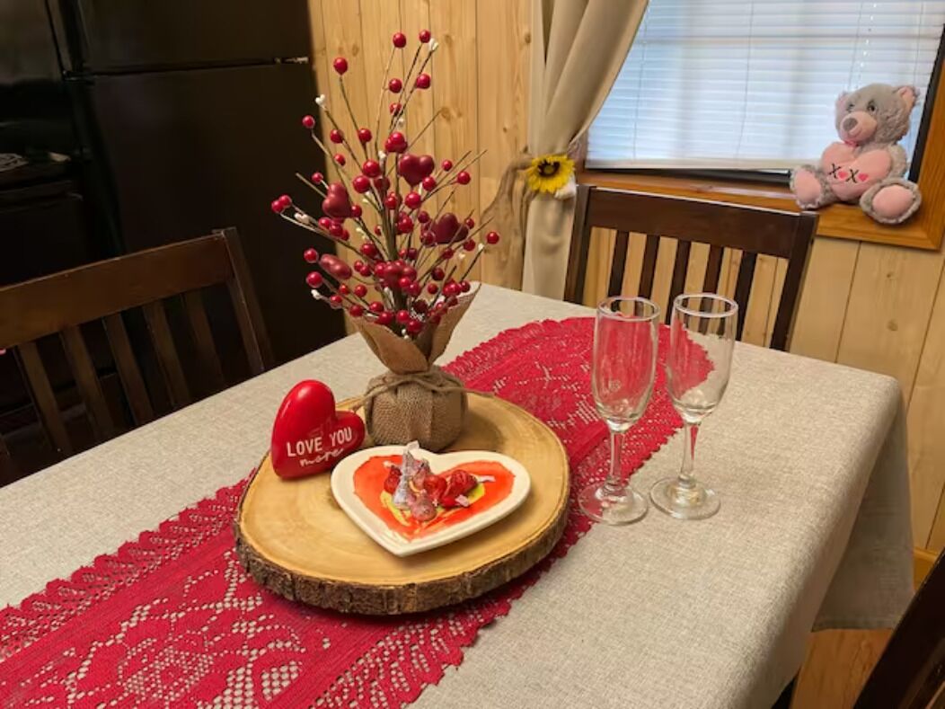 Et spisebord dækket med en rød blonde dug, et træfad med røde bær, en hjerteformet tallerken med mad, en hjerteformet pynteting med teksten "LOVE YOU" og to tomme vinglas.