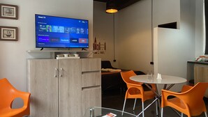 Una camera moderna con TV a schermo piatto che mostra un'interfaccia smart TV, mobili in legno, poltrone arancioni, un tavolino in vetro e una lavagna a parete.