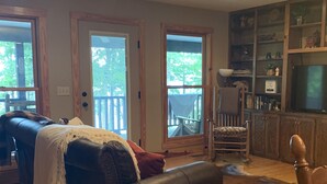 A living room with a dark leather sofa, wooden dining table, and a wooden chair. There is a bookshelf with various items and a television. - 5⭐️ Breathtaking Bluff Views Serene 2 Bed/2Bath Brow Cabin Cloudland near Mentone (Cloudland)