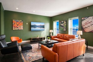 Een woonkamer met groene wanden, een zwarte leren bank, een oranje leren bank, een zwarte fauteuil, een salontafel en een televisie.