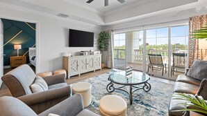 A spacious living room with a ceiling fan, a glass coffee table, and a view of the outdoors through large sliding glass doors. - Beautiful Condo with Golf Course Views, Near Attractions (Kissimmee)