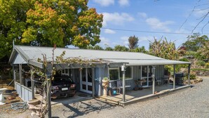 A house with a covered porch, a car parked outside, and a garden area with plants and a barbecue. - Northern Lodge - cottage in a quiet rural setting, lush tropical gardens (Kerikeri)
