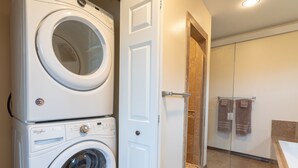 A compact laundry area with stacked washers and dryers, a glass shower enclosure, and a visible bathroom with a bathtub and towels. - Spotted Moon Ranch - River Front - Two Bedroom Cabin (Etna)
