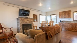 A living room with a stone fireplace, a television mounted above it, and a dining area with wooden chairs and a table. - Spotted Moon Ranch - River Front - Two Bedroom Cabin (Etna)