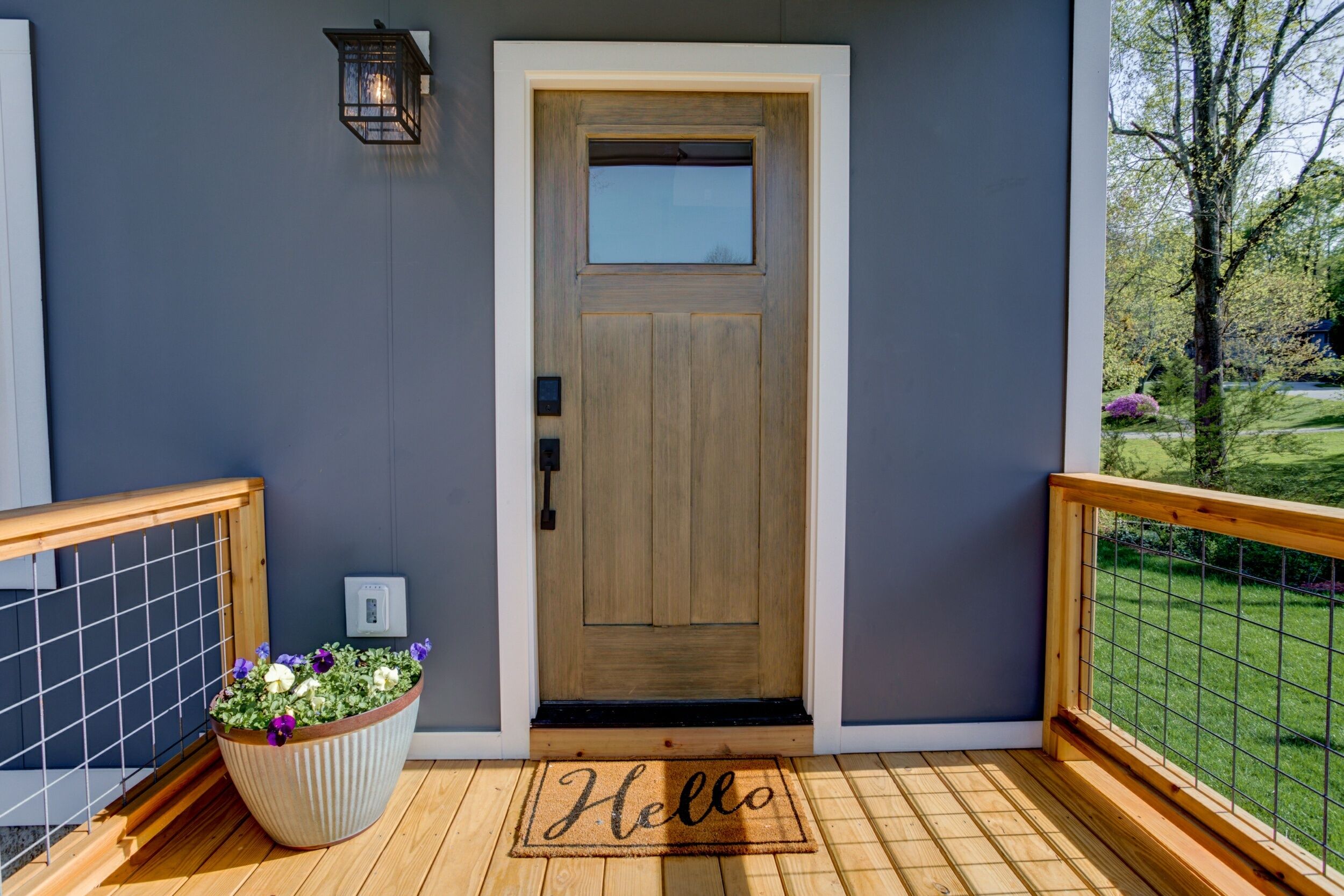 Une porte en bois avec une fenêtre, un pot de fleurs contenant des fleurs violettes et une terrasse en bois sur laquelle est posé un paillasson avec le mot « Bonjour ».