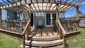 Una terraza de madera con una estructura de pérgola, un espacio cubierto para sentarse y vistas a una casa de color azul.