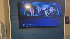 Sinisen seinän päällä on televisionäyttö, jossa näkyy ohjelma. Television alla on takka, jossa näkyy avotuli.