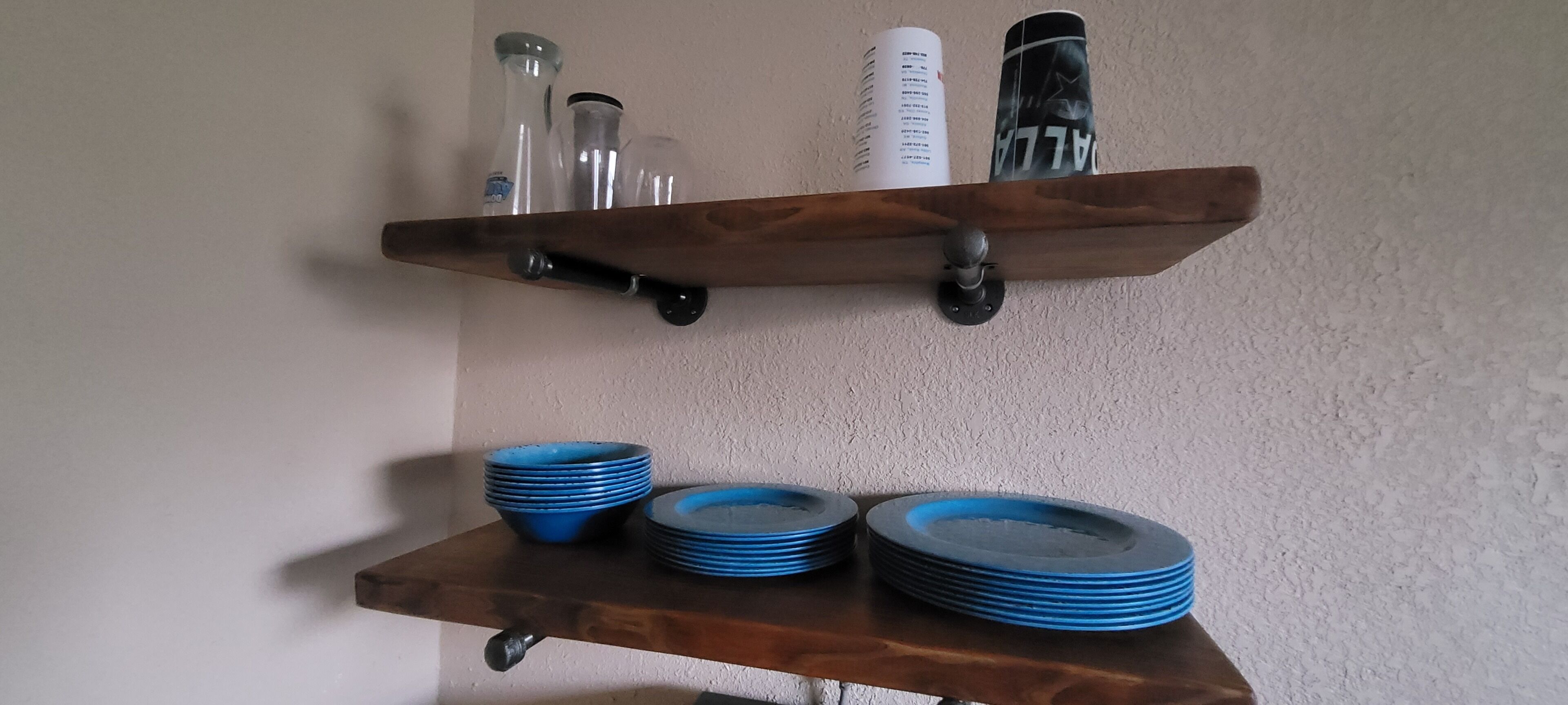 Une étagère en bois avec des bouteilles en verre et un récipient blanc. En dessous, on peut voir des piles d’assiettes bleues.
