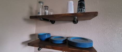 Une étagère en bois avec des bouteilles en verre et un récipient blanc. En dessous, on peut voir des piles d’assiettes bleues.