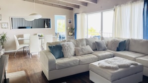 A modern living room with a white sectional sofa, a dining area with a glass table, and a kitchen in the background. - Beach house with unforgettable view (Los Angeles)