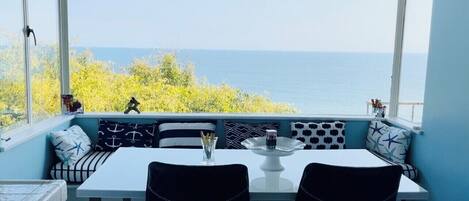 A dining area with a table and chairs, a view of the sea, and a window with a view of trees.