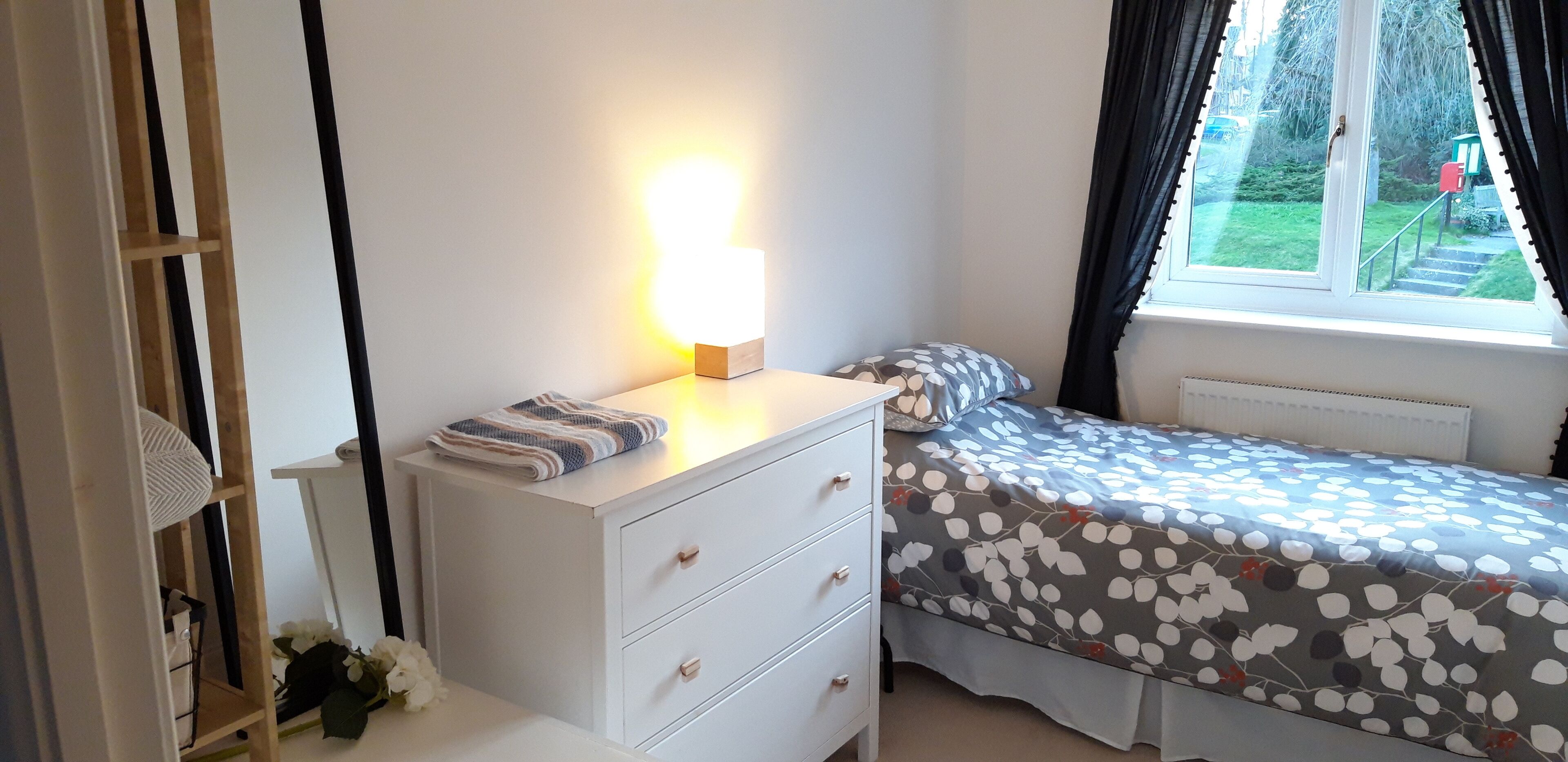 Ein Schlafzimmer mit einem Bett, einem Nachttisch mit Lampe, einer Kommode und einem Fenster mit Vorhängen.