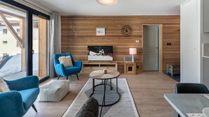 A modern living room with a large TV, a blue armchair, a round coffee table, and wooden paneling on the walls. - Superb 88m2 flat, 3 bedrooms, 2 bathrooms, at the foot of the pistes (Châtel)
