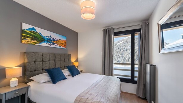 A bedroom with a large bed, a nightstand, a wall-mounted painting, a window with curtains, and a radiator. - Superb 88m2 flat, 3 bedrooms, 2 bathrooms, at the foot of the pistes (Châtel)