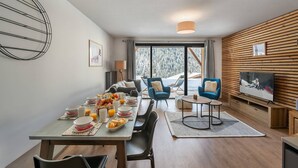 A modern dining area with a wooden table set for breakfast, a TV on the wall, and a view of a snowy landscape through large windows. - Superb 88m2 flat, 3 bedrooms, 2 bathrooms, at the foot of the pistes (Châtel)