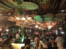A crowded indoor dining area with hanging woven lamps and a central green-lit column.