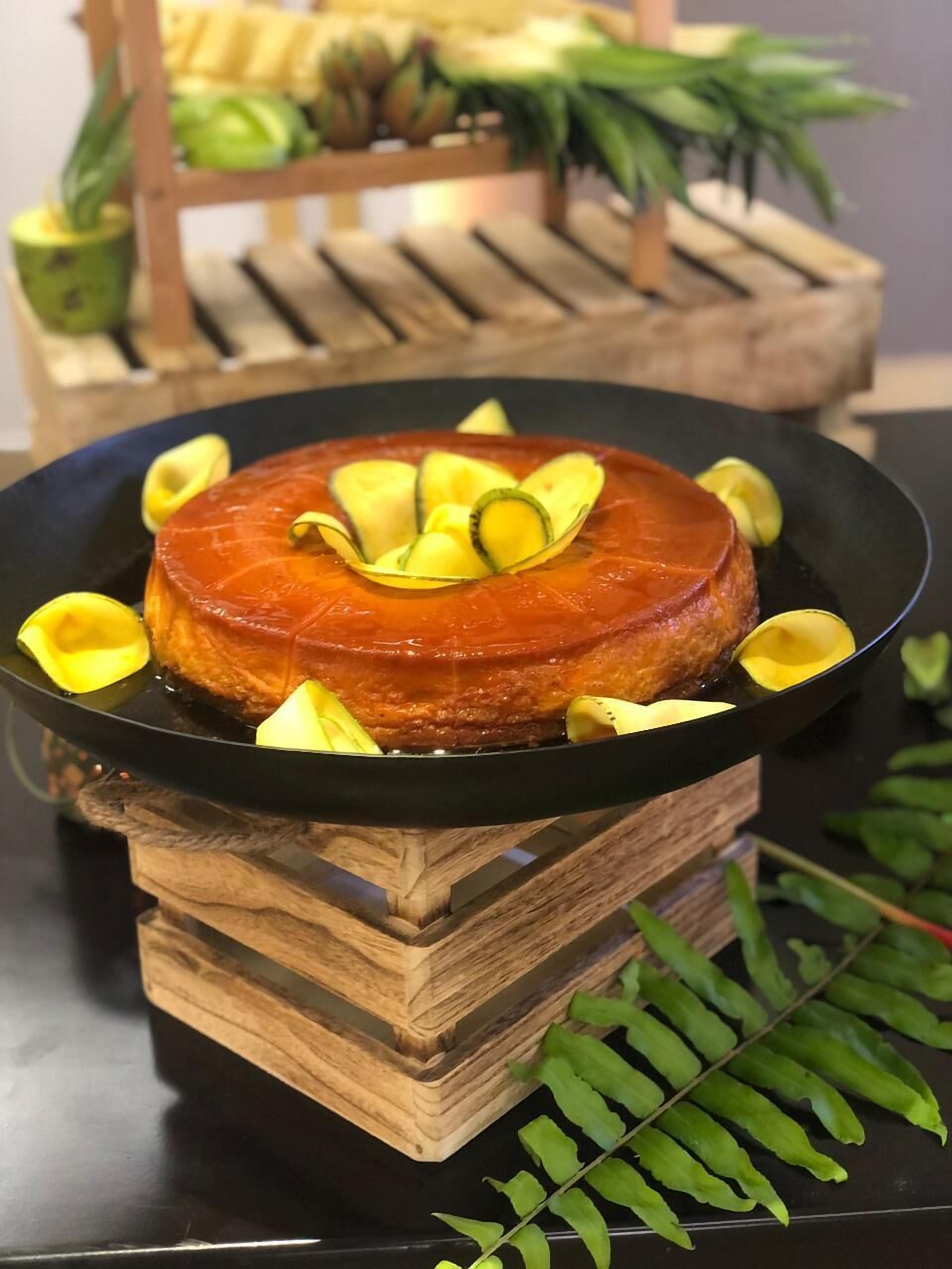 A round, glazed dessert on a black plate, surrounded by yellow fruit slices, placed on a wooden stand.