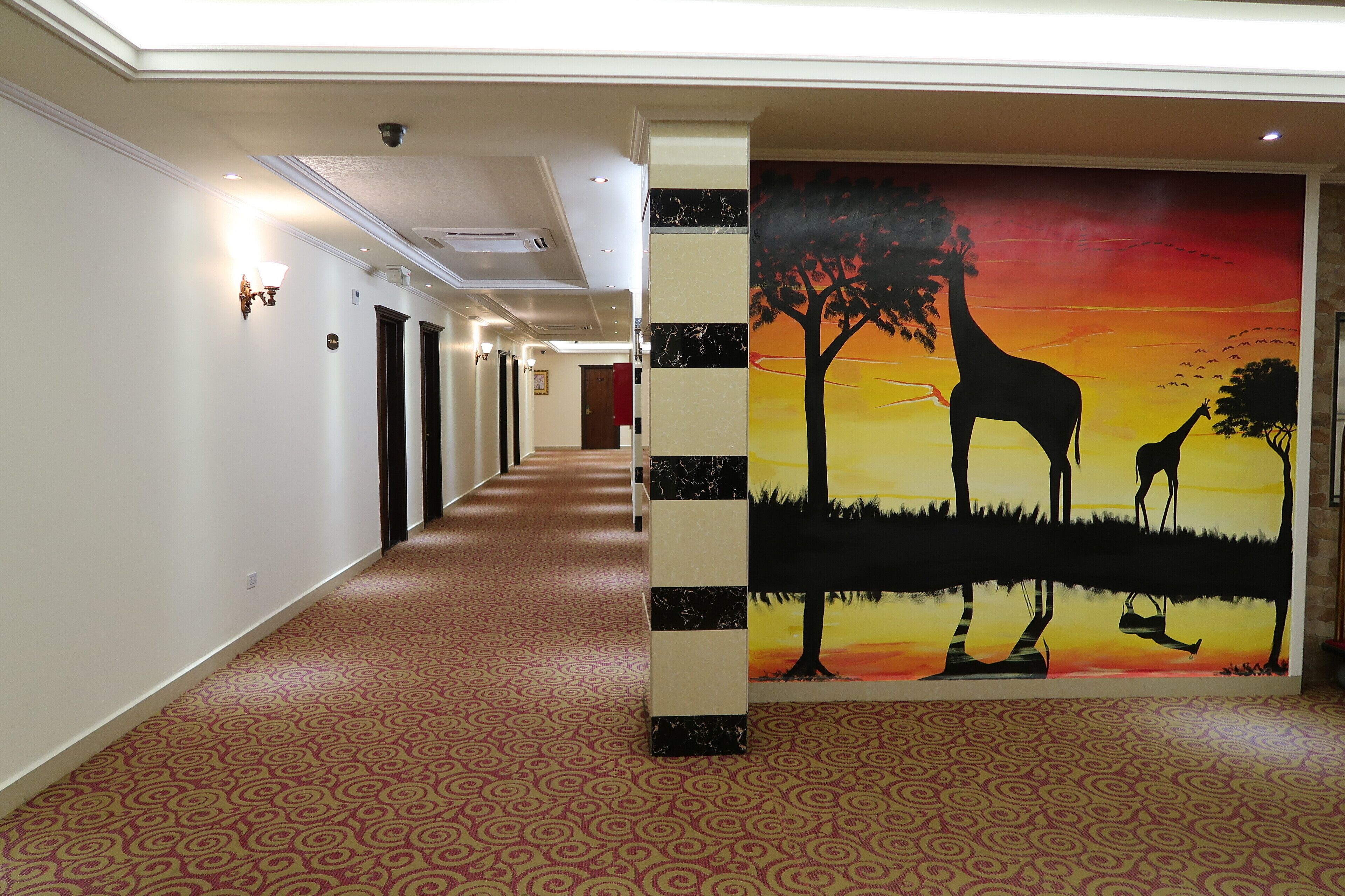 A hotel corridor with patterned carpet, a mural of giraffes and trees, and a row of doors.