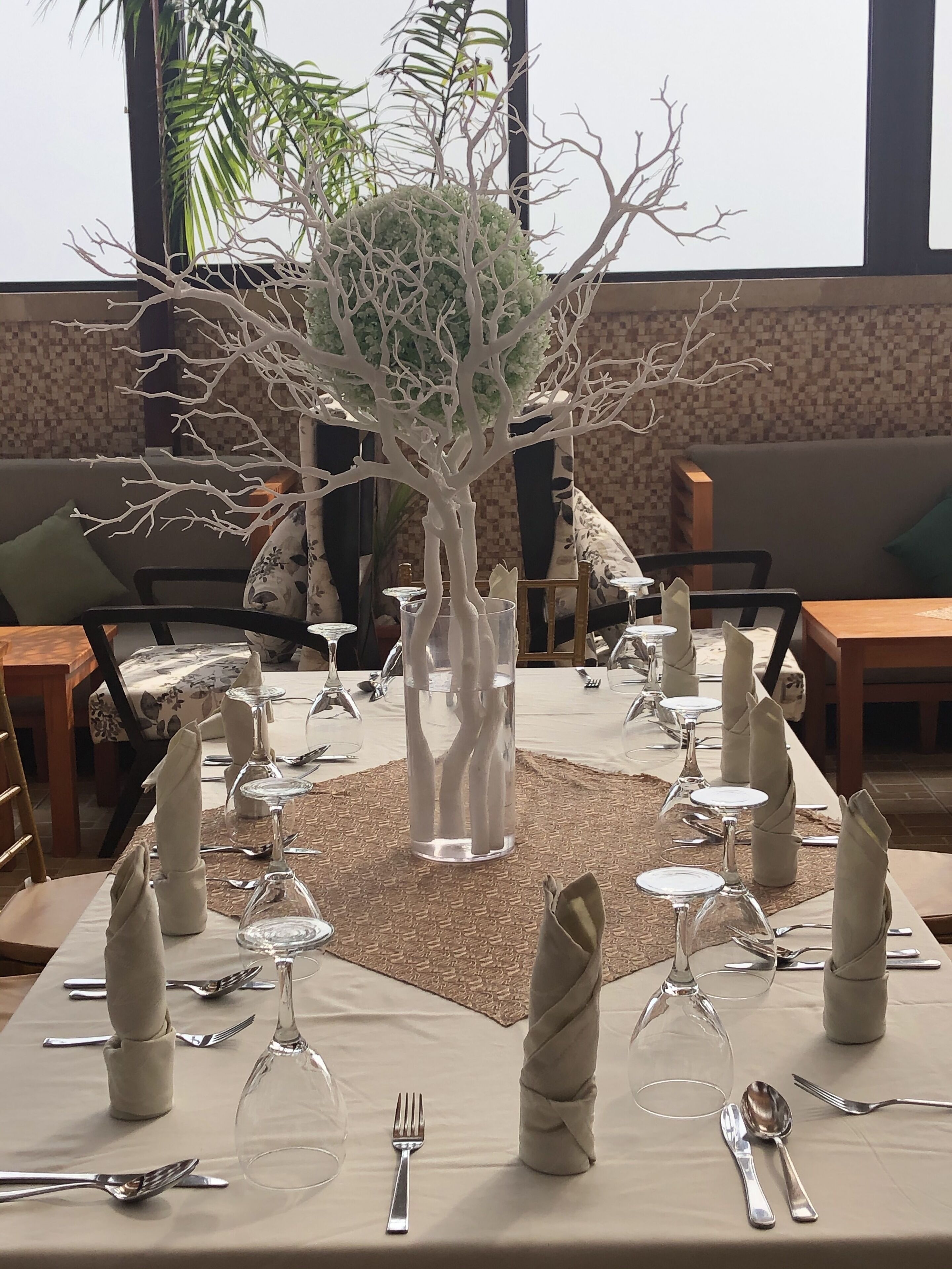 Un espace repas avec une longue table dressée pour un repas, agrémenté de beaux verres, de serviettes pliées et d’un arbre décoratif servant de centre de table.