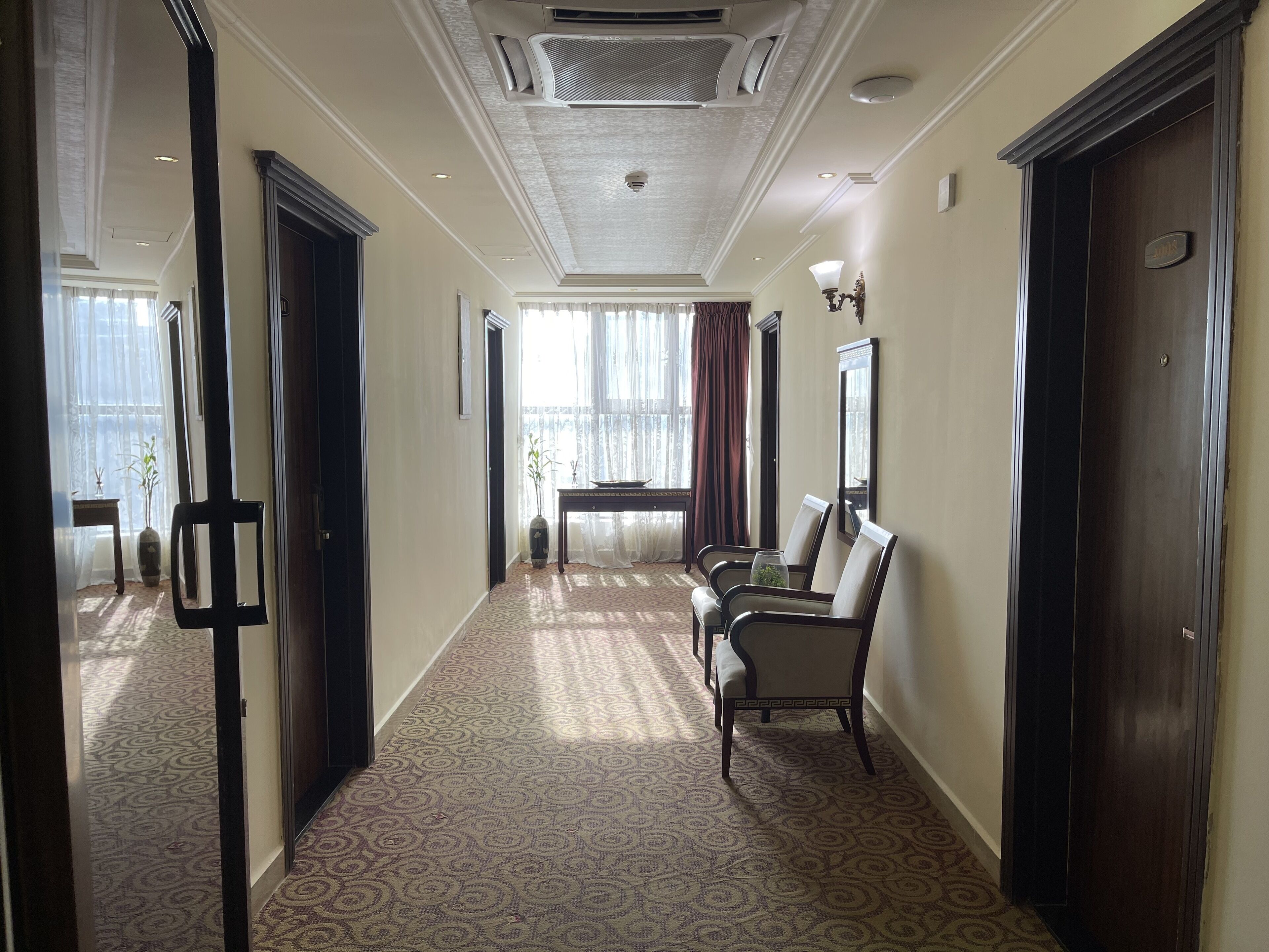 Un couloir d’hôtel avec un tapis à motifs, un coin salon avec des chaises, une table et un grand miroir au mur.