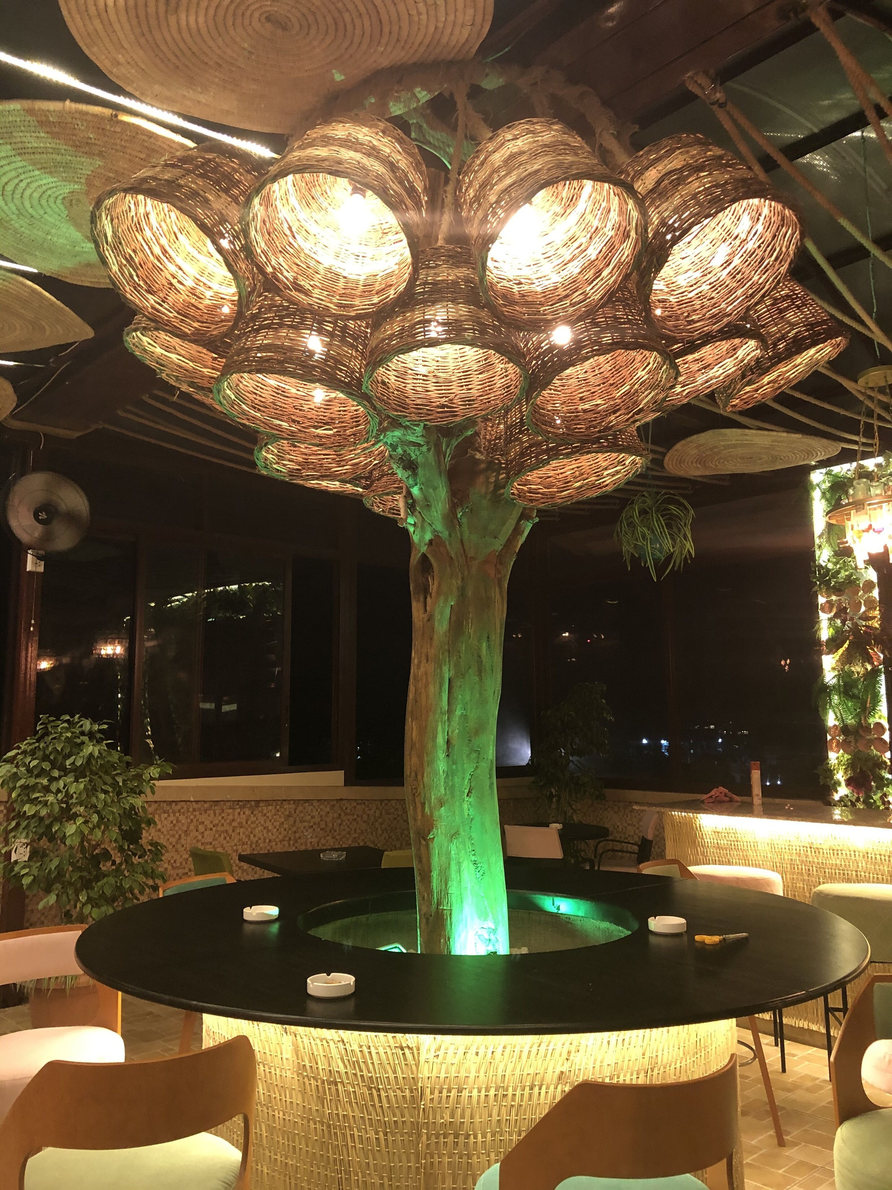 Uma área de refeições com uma mesa central rodeada por cadeiras, uma estrutura de árvore decorativa com luzes penduradas e vista para o céu noturno através de grandes janelas.