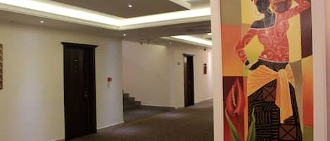 Un pasillo de hotel con alfombra estampada, puertas oscuras y un cuadro colorido en la pared.