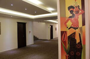 Un pasillo de hotel con alfombra estampada, puertas oscuras y un cuadro colorido en la pared.