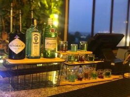 Una zona de bar con botellas de ginebra Hendrick's, Bombay Sapphire y Tanqueray, junto con varios vasos para chupitos que contienen líquidos de distintos colores.