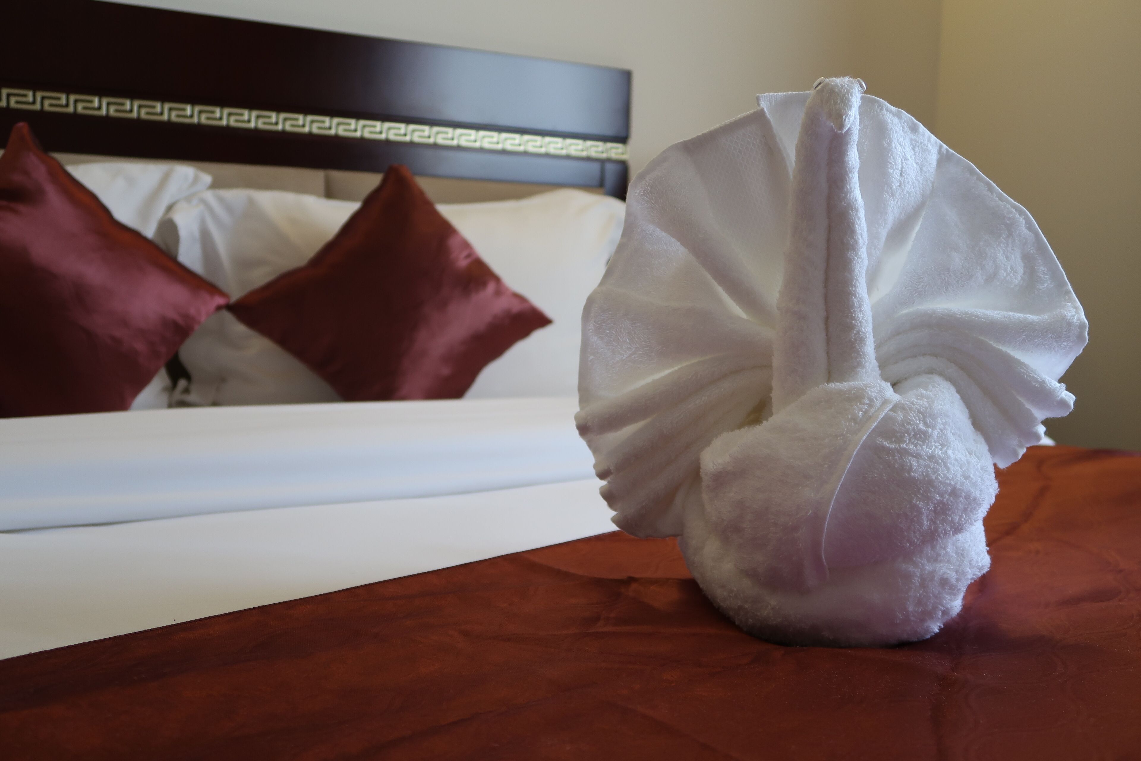 A neatly made bed with white linens and two red pillows, a white towel, and a wooden headboard with a decorative pattern.
