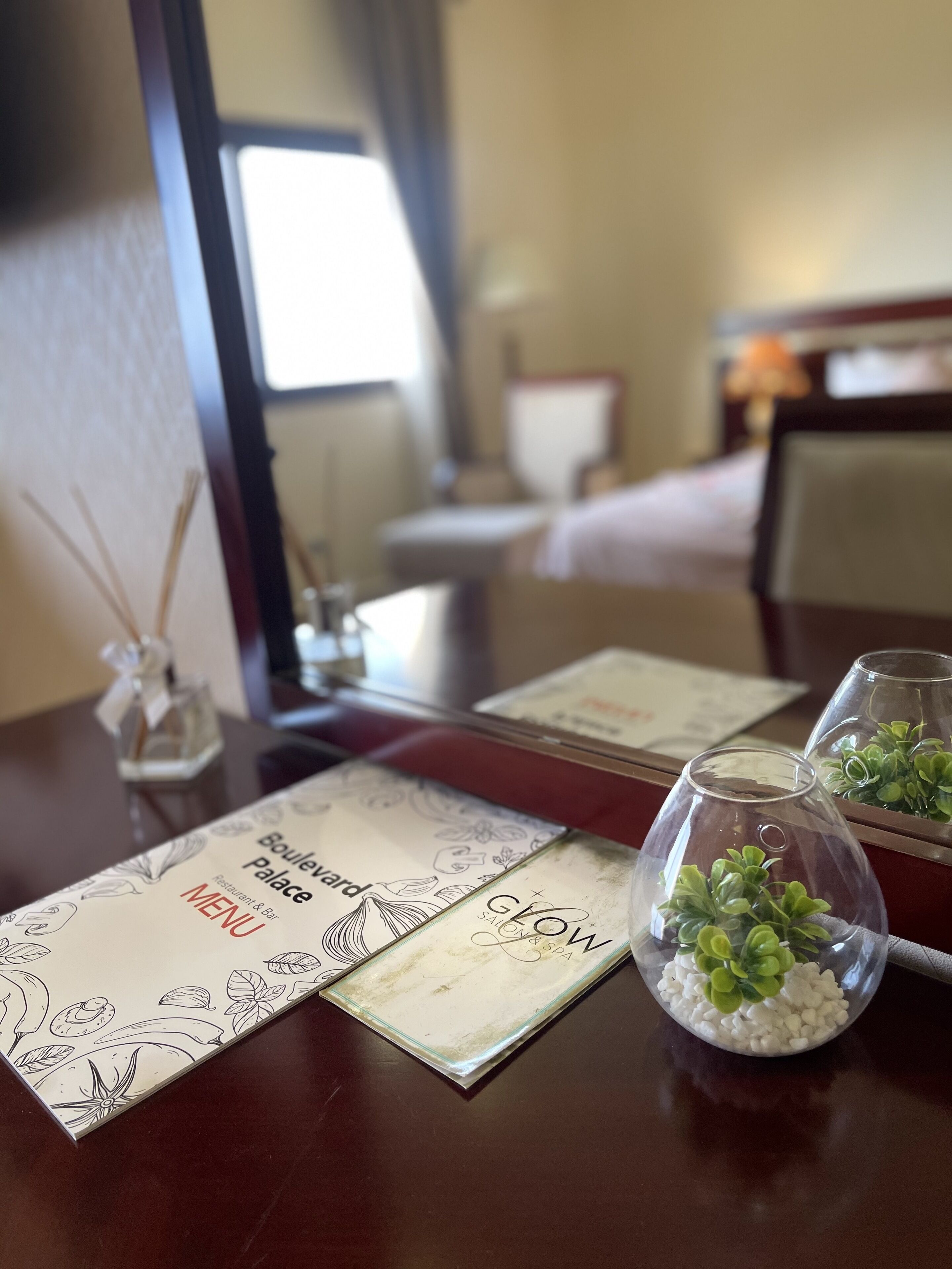 A hotel room with a dining table set with menus, a vase with green plants, and a mirror reflecting the room.