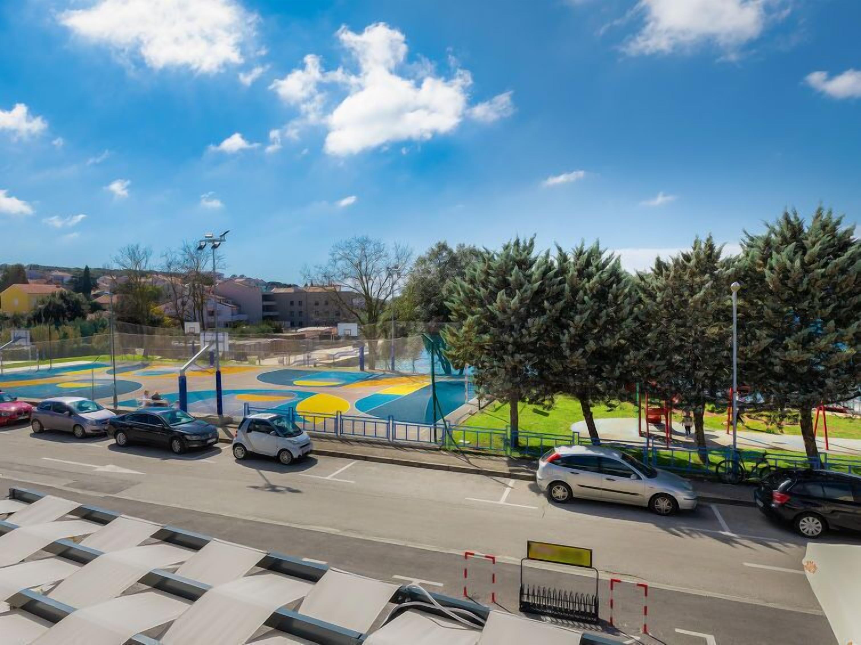 Un estacionamiento con coches, un parque infantil con diseños coloridos, y una carretera con árboles y edificios al fondo.
