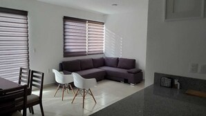 A modern living room with a sectional sofa, a dining table with chairs, and large windows with blinds. - 58th Ave. Apartment (Playa del Carmen)